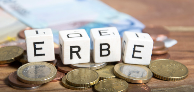 Weiße Buchstabenwürfel, die das Wort "Erbe" bilden; auf Münzen, im Hintergrund Geldscheine; SHBB Bad Oldesloe