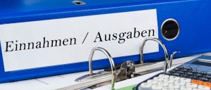 Aktenordner auch Schreibtisch liegend, auf dem Ordner-Rücken steht Einnahmen/Ausgaben, rechts vorne ein Taschenrechner