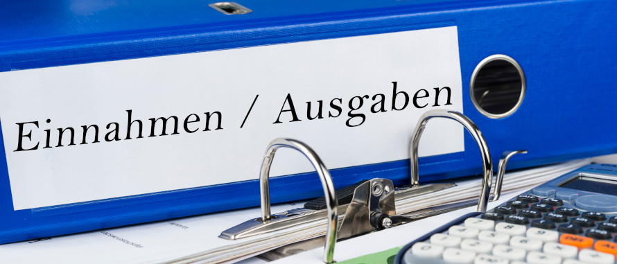 Aktenordner auch Schreibtisch liegend, auf dem Ordner-Rücken steht Einnahmen/Ausgaben, rechts vorne ein Taschenrechner