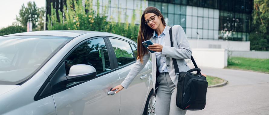 Frau im Business-Anzug, dunkle , lange Haare, blickt auf ein Smartphone und öffnet gleichzeitig die Tür Ihres silberfarbigen Autos